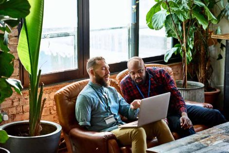 Two men on a leather sofa in a plant-filled room, looking at a laptop together
