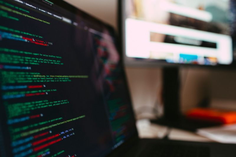Close-up of a laptop screen showing syntax-highlighted code in an editor, second monitor behind